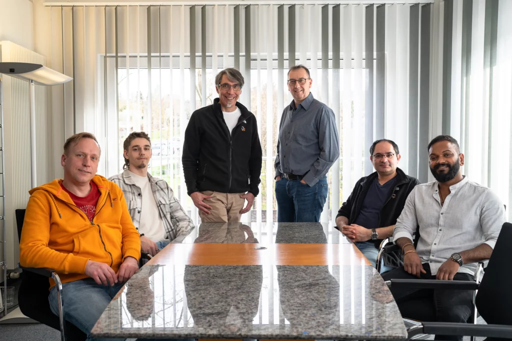 Das Team der DT Netsolution sitzt um einen Tisch und schaut in die Kamera.