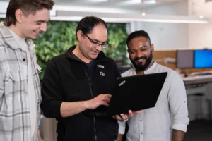Ein Mitarbeiter zeigt seinen beiden Kollegen etwas auf einem Laptop.